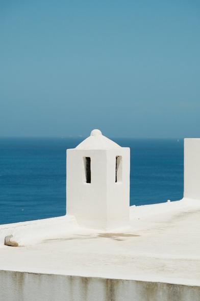 a minimalist picture of a plastered white chimney top with small apertures, topped by a cupola. the blue sea meets the sky in the background