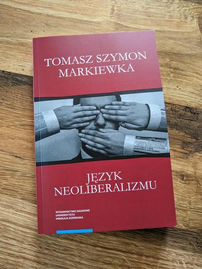 Książka „Język neoliberalizmu” Tomasza Szymona Markiewki leży na drewnianym stole. W centrum jest zdjęcie osoby z oczami i ustami zasłoniętymi dłońmi wystającymi z rękawów z gazet. Nad zdjęciem na ciemnoczerwonym tle imiona i nazwisko autora, pod tutuł. W lewym dolnym rogu nazwa wydawnictwa: Wydawnictwo Naukowe Uniwersytetu Mikołaja Kopernika.