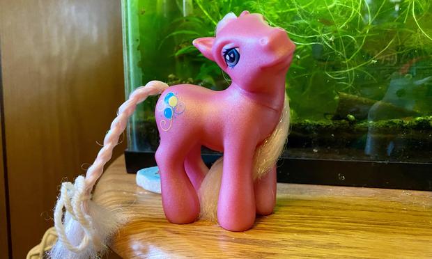 A pink toy pony figurine with a flowing mane and a balloon design on its side, set against a backdrop of green aquatic plants.