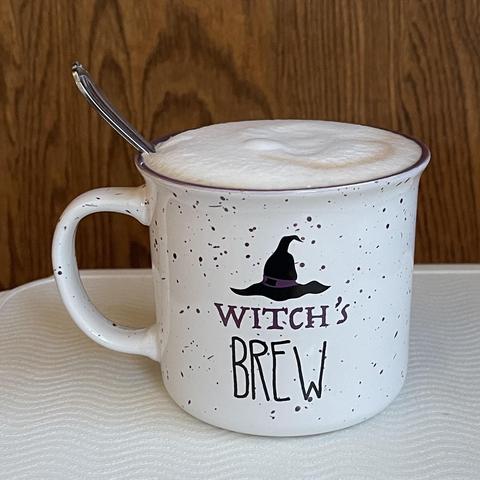 Photo of a ceramic mug in the shape of an enamel tin, off-white with purple speckles and filled to the top with foam. cartoon of a black witches hat with a purple band and the text “witch’s brew.”