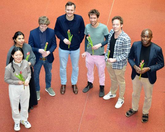 Seven graduates are standing in a half-circle, holding tulips in their hands.