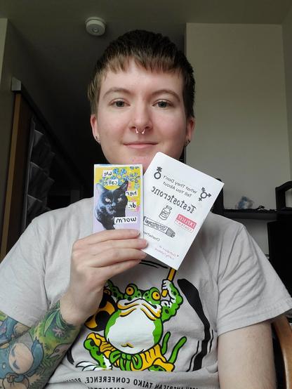 Charlie is a white person with short brown hair and a white tee shirt with a frog on the front. They hold up two zines facing the camera. The mini zine features a photo of their black cat on the cover. The quarter page sized zine features testosterone doodles.
