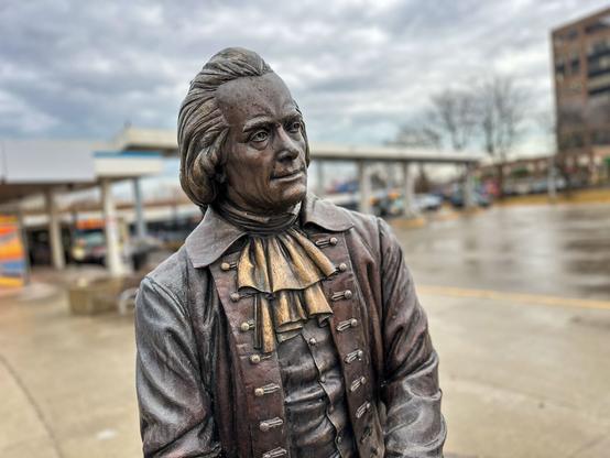 A bronze statue of a historical figure, dressed in an 18th-century outfit, featuring a ruffled collar and a jacket. The background includes a cloudy sky and a parking lot with vehicles.