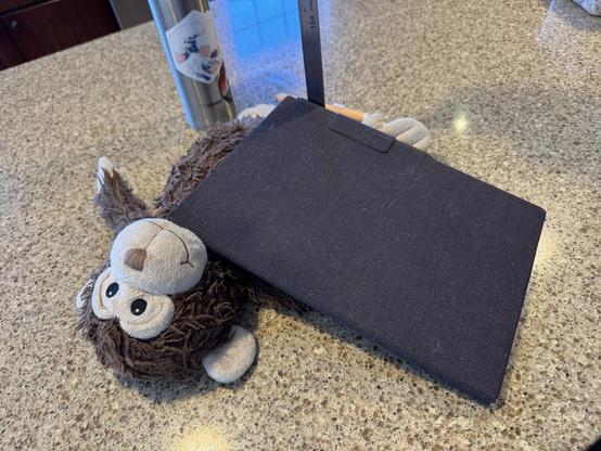 A reMarkable paper pro in it’s official case . It is leaning on a stuffed monkey on a granite countertop. There is a tumbler full of water in the background.