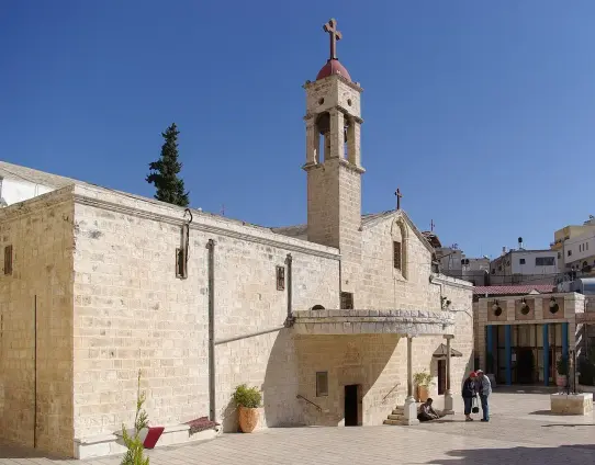 The image features a stone-built church with a bell tower topped by a cross. The architecture appears traditional, with a clear blue sky in the background. There are a couple of people visible in front of the church, and the surrounding area seems to include some greenery and modern buildings in the distance. The overall setting looks peaceful and inviting.

Image Credits: Wikimedia / Berthold Werner / Public domain