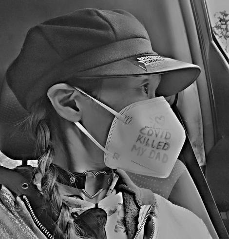 A black & white selfie photo of me wearing a white N95 face mask with the handwritten message "COVID KILLED MY DAD" accompanied by a small heart drawn above it. I'm wearing a black hat with a sticker that says "BOYCOTT ISRAEL APARTHEID". A braid is visible on the side of my face. I'm sitting in side a car and looking out through the car window. The background is blurred, implying movement of car. I'm wearing a black leather choker with a hollow metal heart centered.