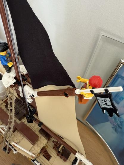 A close-up of a LEGO pirate ship featuring a pirate figure waving a black flag with a skull and crossbones. The ship has a beige sail and various details, including a second pirate figure visible on deck. The background shows a blurred photo frame.