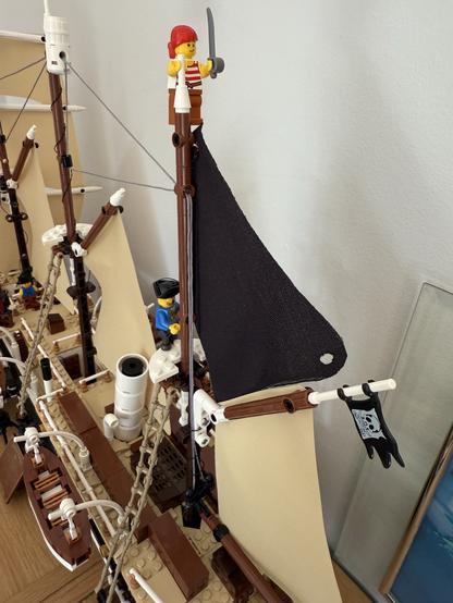 A detailed LEGO pirate ship model featuring a pirate minifigure holding a sword atop the mast. Another minifigure stands on the deck, and there are sails and a pirate flag visible. The ship's design includes various brick elements and rigging.