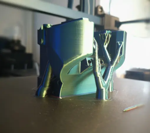 eine grünlich schimmernde Figur die von fein verzweigten "Baumstützen" supportet wird.
Das Objekt steht auf dem Druckbett und im Hintergrund ist ein Fenster und noch ein bisschen  was vom 3D Drucker zu erkennen.