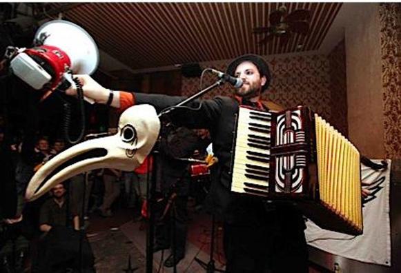 A whitefaced bearded man in a hat, wearing a piano accordion, gestures with a megaphone before a mic stand with a bird mask hanging on it.