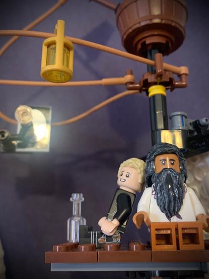 Lego Ed in a white shirt and copper pants and Lego Stede is black leathers leaning against the mast. A lantern, mirror, and spyglass hang above them.