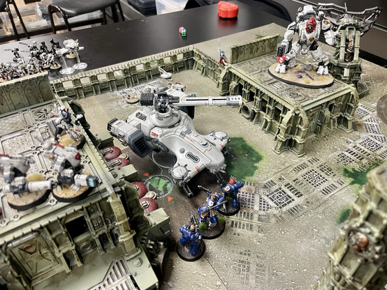 A t’au hammerhead tank between some bunkers with a ghostkeel standing on the roof of one bunker and stealth battlesuits on another. Facing them is an ultramarines intercessor squad.