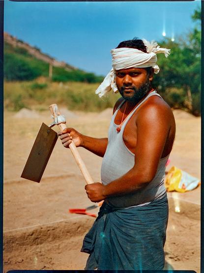 Color photo of a man holding a hoe with an excavation area in the background.