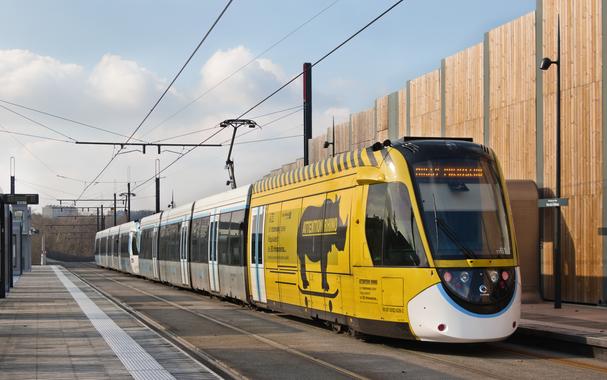 Une rame de tram-train de type U 52600 (ou Alstom Citadis Dualis), de la ligne 12 du tramway d’Île-de-France, à l’arrêt Parc du Château. La queue de la rame, au premier plan, a un pelliculage jaune sur toute la première voiture avec le message « Attention rhino » et un dessin de rhinocéros sur une planche à roulettes. Le reste de la rame est dotée de la livrée blanche, bleue et grise caractéristique d’Île-de-France Mobilités.