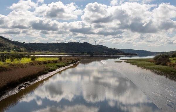 View of the Guadiana river on the way to Alcoutim (Portugal). At the bottom of the photo there is water across the entire width, in the distance the river bends to the right. The sky with many white and gray clouds reflects in the water. In the distance mountains.