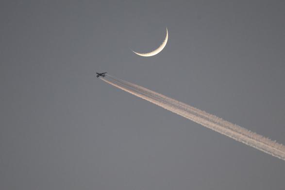 Der Mond als Sichel (weiß) darunter ein Flugzeug mit einem grossen Kondensstreifen am abendlichen Himmel.
Marzework