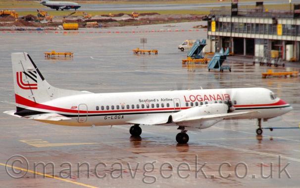 Side view of a white, twin propellor-engined airliner being pushed back from it's stand by an out-of-frame tug connected to the nose-wheel by a long, thin blue pole.
There is a black and red stripe running along the body. With red "LoganAir" titles on the upper forward fuselage, and smaller black "Scotland's Airline" titles on the upper rear.
The registration "G-LOGA"  is on the lower rear fuselage in black.
The tail has a stylised red upper-case "L" with an arrowhead pointing to the upper right.
Wet concrete apron fills most of the frame, with a terminal building coming in from the right side, and a grey and dark blue jet airliner landing on a runway in the distance.