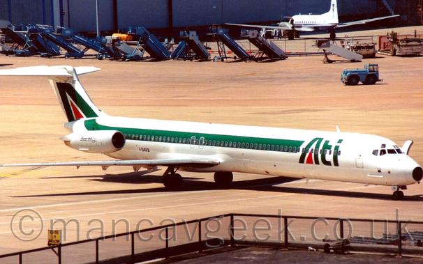 Side view of a white, twin engined jet airliner with the engines mounted on the sides of the rear fuselage, parked facing to the right.
The plane has a green stripe running along the body and sweeping up into the tail, where it forms the outline of an upper-case letter "A", with a red triangle as the centre part.
There are large green "ATI" titles on the forward fuselage, again with a red triangle in the centre of the "A".
The registration "I-DAVB" is on the lower rear fuselage in black, and the letters "VB" on the front of the cockpit roof, also in black.
The forward door on the opposite side is open, indicating that the plane is parked, even though it is nowhere near the terminal, and in fact is blocking a roadway marked on the concrete apron, which runs directly under the fuselage.
The roof of part of the terminal can be seen in the foreground, with a black metal fence around the edge.
Sets of blue mobile airstairs are lined up in the background, in front of a large grey hangar.
Parked in front of the hangar is a white and blue twin propellor-engined airliner.