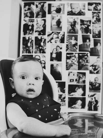 Image of a baby in highchair. Behind him, there’s a side of a fridge with printed photos on it. The baby is looking at the camera.