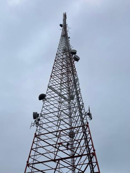 A red and white open framework broadcast tower with a triangular cross section extends up the middle of the image in front of a grey cloudy sky. Various smaller antennas and dishes hang off of it.