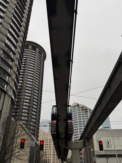 Two monorail tracks against a grey sky above the viewer. The left track is centered in the image with the red and white train traveling down and into the distance towards other office towers. To the left are two angled concrete brutalist round hotel towers. In the foreground are a set of red stop lights for each road lane.