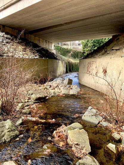 A small stream running through the rocky terrain is transitioning to a concrete walled watercourse under the bridge.