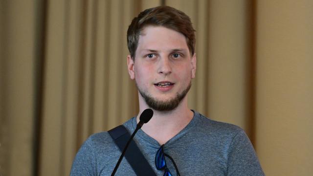 Nahaufnahme von Jonas Will mit kurzen, hellbraunen Haaren und einem kurzen Bart. Er trägt ein hellgraues T-Shirt und spricht in ein Mikrofon, das an einem Ständer befestigt ist. Der Hintergrund ist unscharf und zeigt einen beigefarbenen Vorhang.
