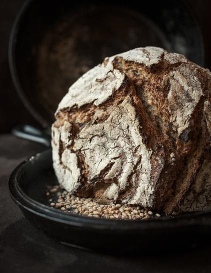 Ein kross gebackenes Roggensauerteigbrot liegt mit dem Anschnitt nach unten in einer Art Pfanne. Vor dem Brot liegen ungemahlene Roggenkörner. Das Licht kommt von links. Der Hintergrund liegt im Dunkeln.