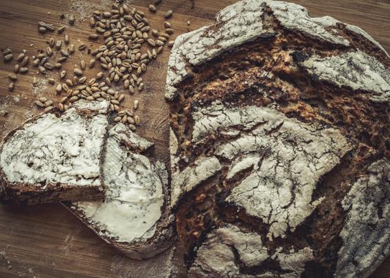 Perspektive von oben: Eine Scheibe Brot, dass in der Mitte auseinandergeschnitten und mit (veganer) Butter bestrichen ist liegt links. In der oberen Ecke liegen lose verteilt Roggenkörner. Rechts sieht man das Brot von oben, dass schön kross aufgesprungen ist. Das Ganze liegt auf einem Holzbrett.