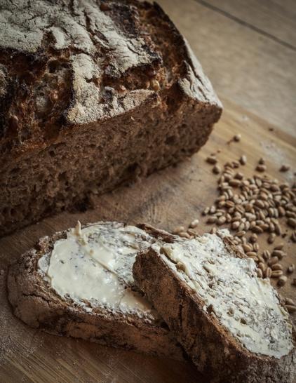 Etwas erhöhte Perspektive: Man sieht das abgeschnittene Brot mit einer Scheibe (in der Mitte auseinandergeschnitten), die mit Butter bestrichen ist. Dahinter liegen Roggenkörner. Das Ganze liegt auf einem Holzbrett.