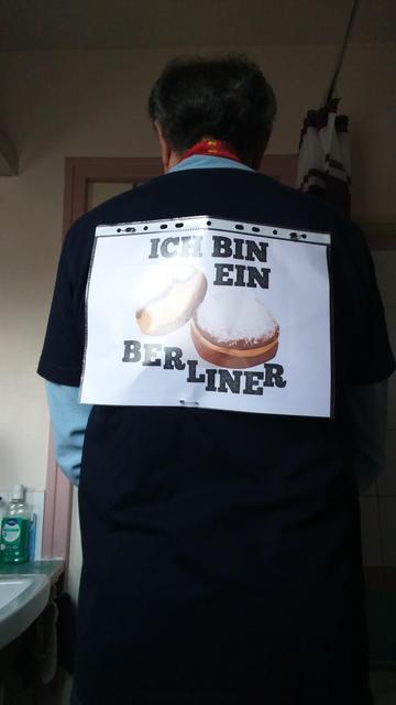 On me voit de dos avec mon panneau accroché à mon T-shirt bleu marine à manches longues enfilé par dessus un sweatshirt bleu ciel à manches longues).

Mon panneau indique "Ich bin ein Berliner", illustré par deux images de beignets.