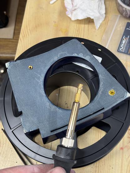 The radar eye of R2-D2 on a filament spool. The back side up. Two holes were drilled into it. A soldering iron with a heat insets is in frame. One inset is already melted into the plastic on the top left of the radar eye.