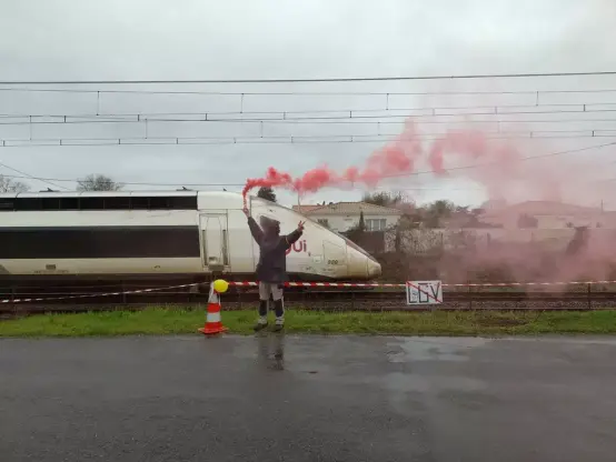 Une personne tien un fumigène le long d’une voie ferrée sur laquelle pace un TGV. Sur une pancarte accrochée la mention « LGV » (barrée) et le dessin d’un poing.