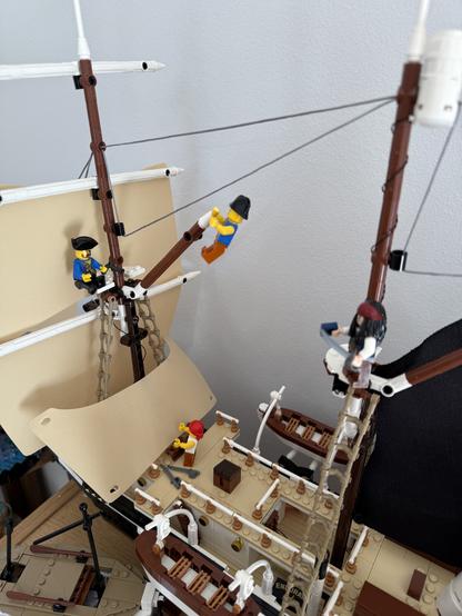 The image features a detailed LEGO ship model with several miniature figures. One figure is climbing the rigging, while others are working on deck, adjusting sails or tending to tasks. The ship has multiple masts, sails, and an intricately designed deck