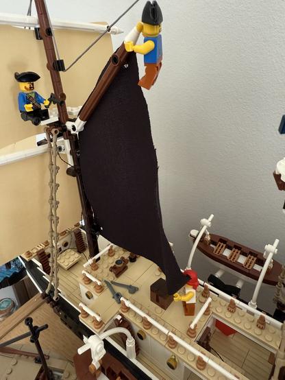 A colorful LEGO ship model with two mini-figures. One figure is climbing to adjust a large black sail, while the other is seated at the ship's wheel. The ship features detailed elements like a deck with barrels, weapons, and other decorations