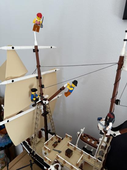 A Lego pirate ship model is shown, featuring several pirate minifigures positioned on the sails and masts. One minifigure stands at the top of the mast, while others are climbing or sitting on the rigging and deck. The ship