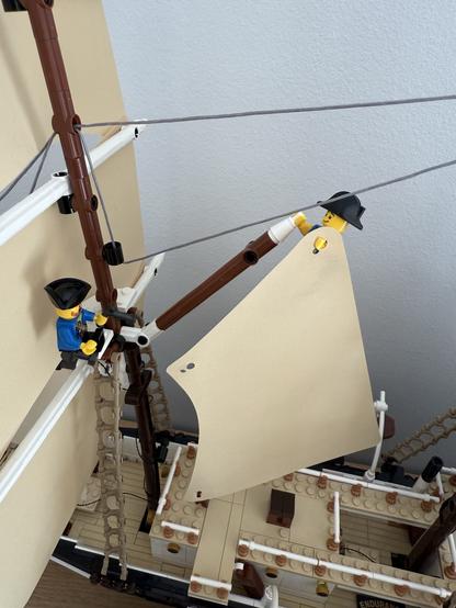A close-up of a model ship made from building blocks, featuring two minifigures. One figure is climbing a ladder, while the other is adjusting a sail. The ship has a detailed deck and rigging, with a plain background.