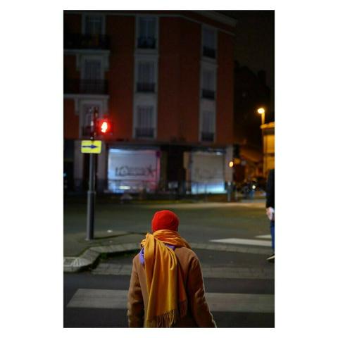 Une personne vêtue d’un manteau beige, coiffée d’un bonnet rouge et enroulée dans une écharpe jaune vif, attend devant un passage piéton. Le feu tricolore rouge illumine discrètement la scène, tandis que l’arrière-plan urbain, avec ses bâtiments et lumières tamisées, crée une atmosphère nocturne calme et contemplative.