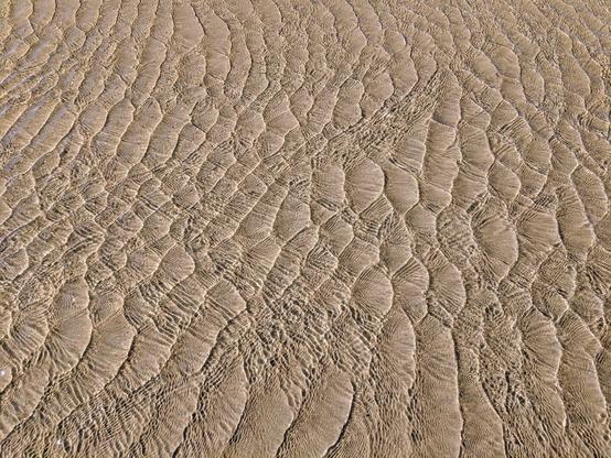 A close-up, overhead shot of shallow water flowing over a sandy surface. The sand is light brown and textured with distinct, repeating ripple patterns, creating a corrugated appearance. The water is clear and reflects light, revealing the sand's texture beneath. The ripples form diagonal lines across the frame, suggesting a gentle current. The overall effect is a natural, abstract pattern of light, water, and sand.