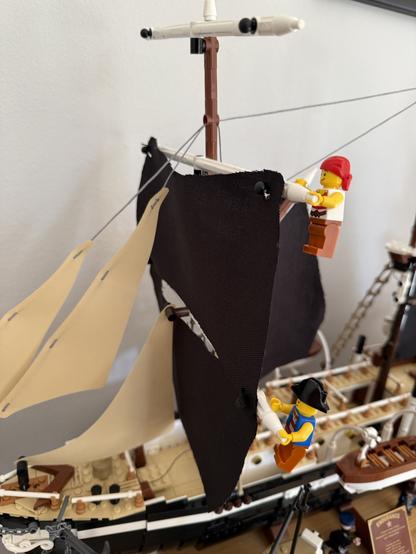 The image features a model ship with two LEGO figures. One figure, wearing a red bandana, is climbing the mast, while the other, dressed in pirate attire, is handling a sail. The ship has multiple sails, and the background includes other