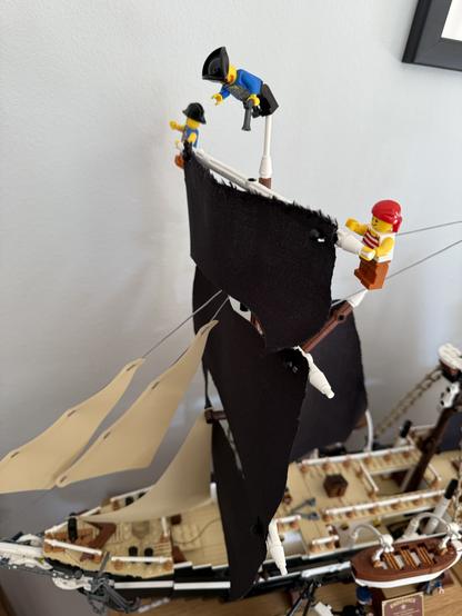 A close-up of a model pirate ship with three Lego minifigures. Two figures are climbing the sails, while one is positioned on the deck. The ship features black and tan sails, intricate details, and is set against a plain wall.