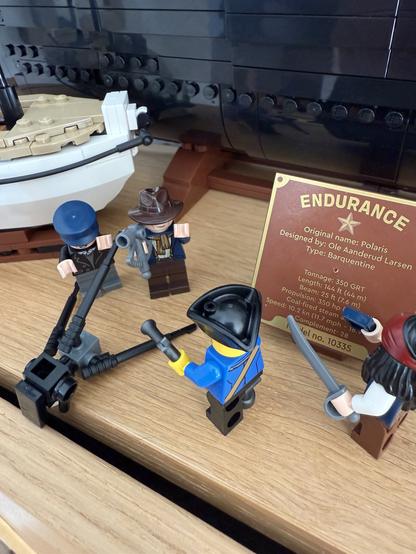 A Lego diorama featuring four mini-figures dressed as sailors or pirates, engaged in various activities near a model of a ship named "Endurance." A plaque detailing the ship's specifications is visible in the background.