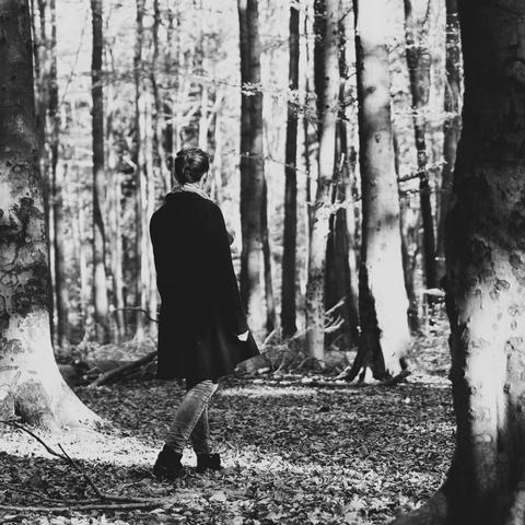 Schwarz-weiß-Foto einer Frau die durch einen Laubwald im Herbst schlendert. Sie ist von hinten zu sehen und trägt einen dunklen Mantel und Jeans