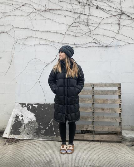 Woman standing in alley, wearing a hat and a long black coat, and with her head turned to the left