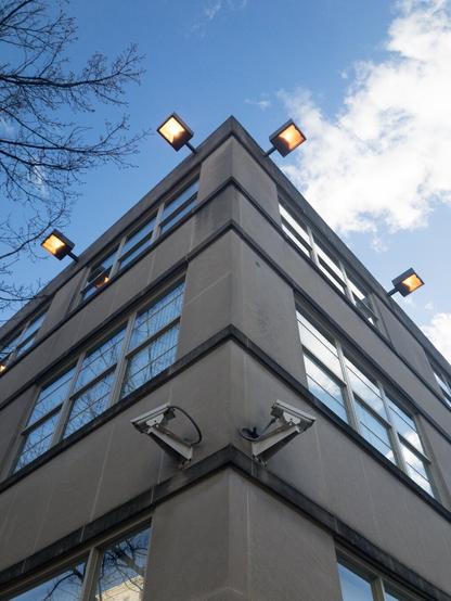 Looking up at the corner of a building with two security cameras and four flood lights turned on during the daytime