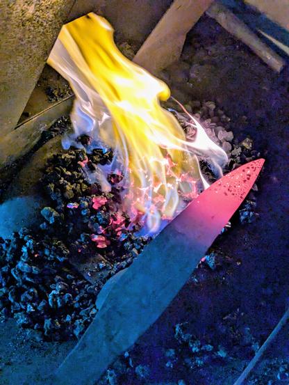 Le foyer de forge avec de drôles de couleurs. Les flammes sont aspirées dans la hotte. Je tiens la lame dont la pointe est rougeoyante au dessus du feu.