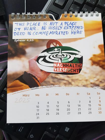 Foto eines Kalenderblattes vom Monat März. Das Bild zeigte ursprünglich einen Fluss im Hintergrund und im Vordergrund eine Hand die einen Kompass hält. Der Kompass wurde mit einem Sticker auf dem "nachhaltig verseucht" steht überklebt. 
Der bibelvers in der linken oberen Ecke wurde überklebt mit dem Text:
"This place is not a place of honor. No highly esteemed deed is commemorated here". Darunter steht noch die Quelle des ursprünglichen bibelvers: Epheser 4.23