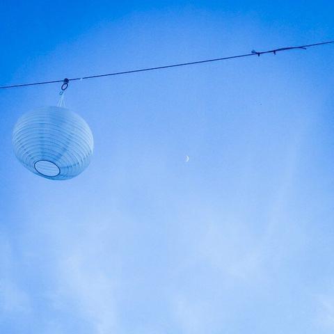 A white lampion hanging on a wire in front of the clear blue sky. You can sense the half-moon in the back.