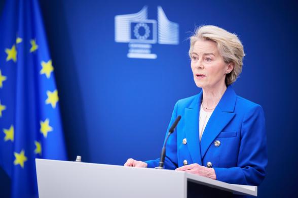 Ursula von der Leyen, President of the European Commission, delivers a speech at a podium. She is wearing a royal blue blazer with gold buttons, and a European Union flag is visible in the background. The backdrop features the European Commission logo on a deep blue background.
