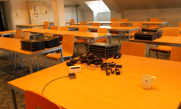 Klassenraum mit Stühlen, Tischen und gestapelten Laptops. Im Hintergrund ein Fenster in einer Dachschräge.